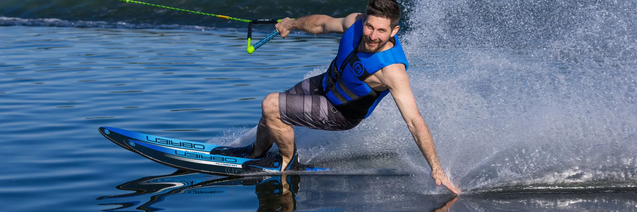 O’Brien 2026 Celebrity 68-inch Blue Combo Waterskis with X-7 Bindings 1 Man water skiing on a lake with a blue ski and life jacket.