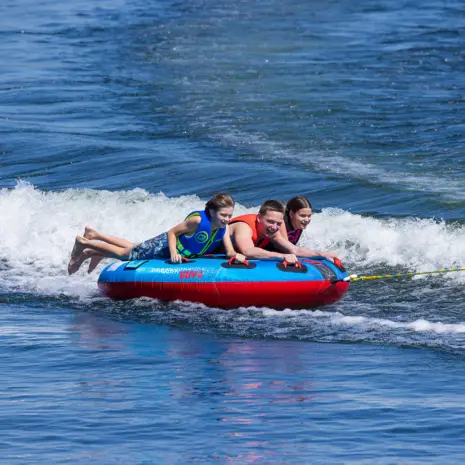 Obrien Ultra Screamer Towable Tube Lifestyle 05 Three people on a red and blue Ultra Screamer towable tube being pulled by a boat on a body of water.