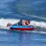 Three people on a red and blue Ultra Screamer towable tube being pulled by a boat on a body of water.