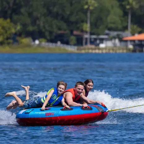 Obrien Ultra Screamer Towable Tube Lifestyle 06 Three people on a blue and red Ultra Screamer tube in the water with a blurred background of trees and houses.